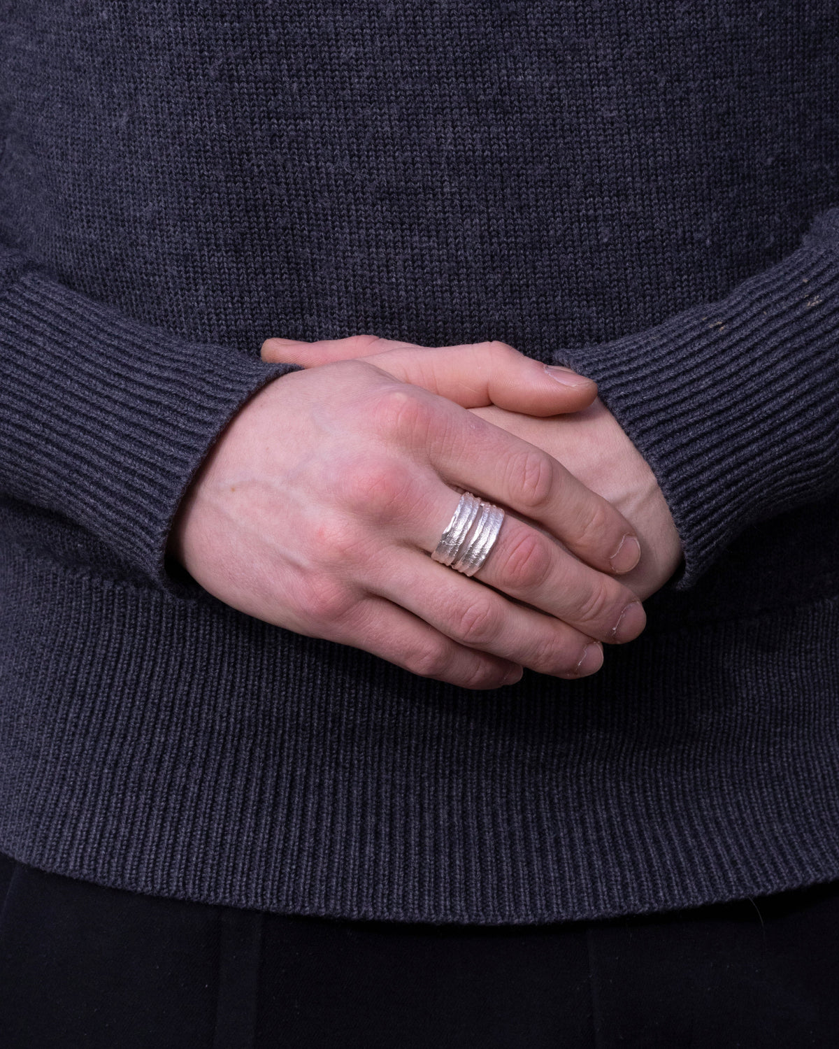 Silberring mit Asphaltstruktur am Finger eines Mannes, Hände vor grauem Strick gefaltet