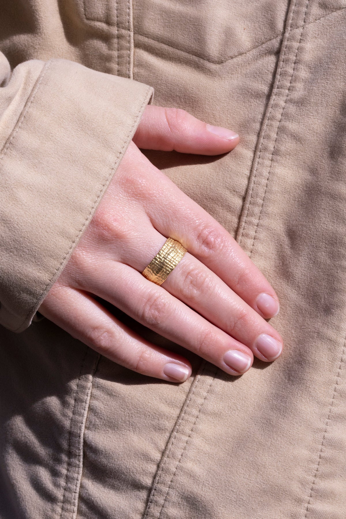 Breiter Ring aus Gelbgold mit fein strukturierter Oberfläche an schlanker Hand, beige Jacke