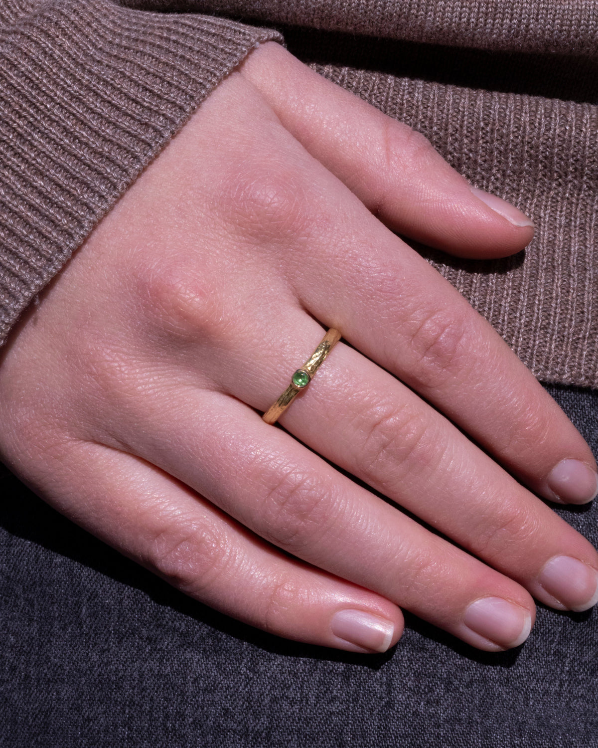 Gelbgoldring mit grünem Tsavorit und Rindenstruktur am Finger einer Frau, Hand auf grauem Stoff