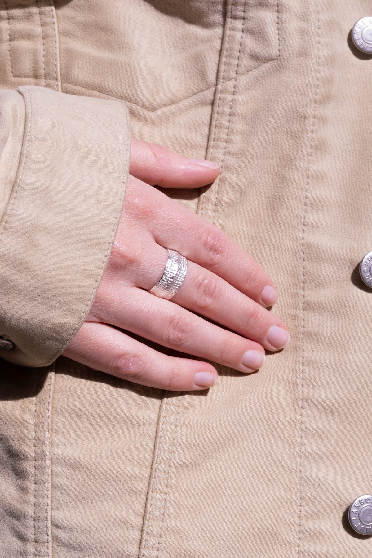 Breiter Silberring mit feiner Nugget-Struktur am Finger, Hand ruht auf beiger Jacke