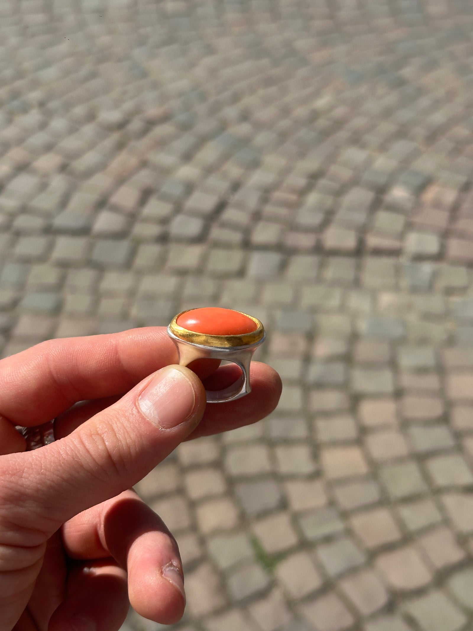 Silberring mit rotem Korallen-Cabochon in Goldfassung, zwischen Fingerspitzen über Kopfsteinpflaster
